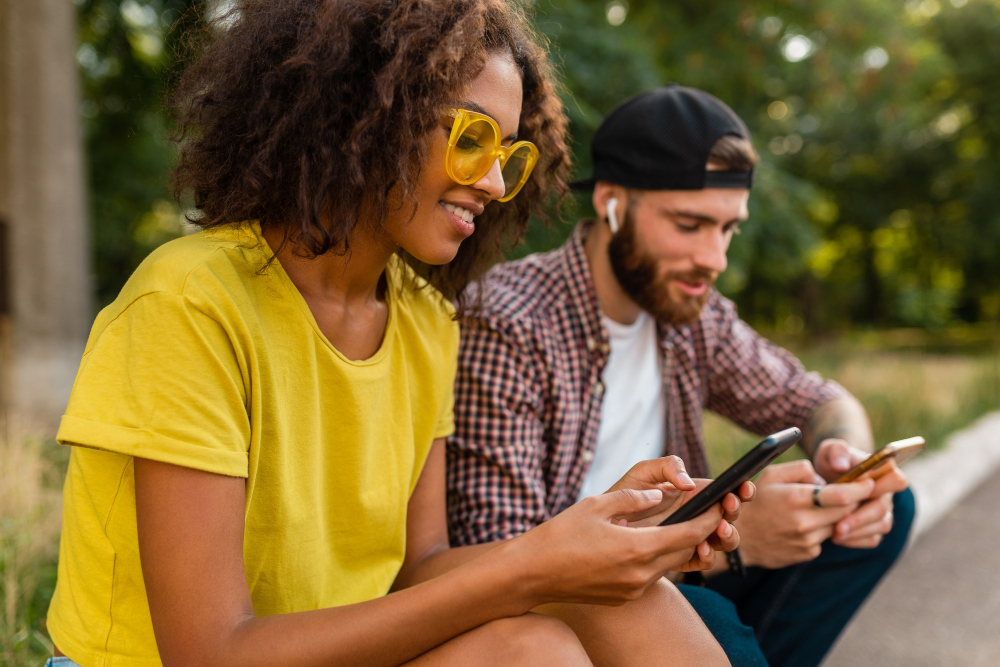 amigos-sonrientes-jovenes-felices-que-sientan-parque-usando-smartphones-hombre-mujer-que-divierten-juntos